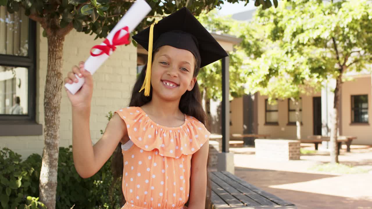 Niña afroamericana de pie en un banco escolar con birrete, celebrando y ondeando un diploma con cinta roja