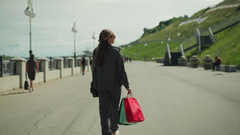 back view of lady in grey clothing holding red, mint and green shopping bags, walking on bridge, light poles line one side with a hill on the other, people strolling ahead in the background