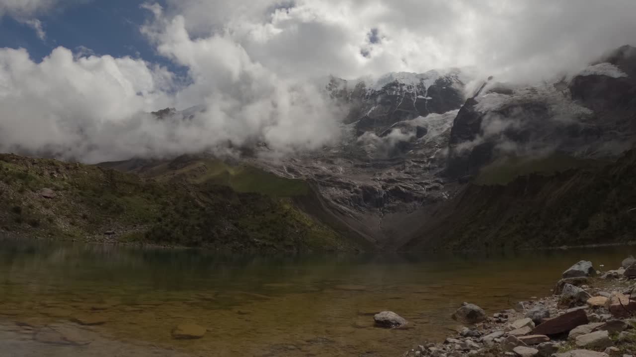timelapse diurno en el lago humantay en perú con nubes moviéndose entre las montañas reflejándose en el agua azul clara con nieve en la parte superior y rocas en primer plano sin gente