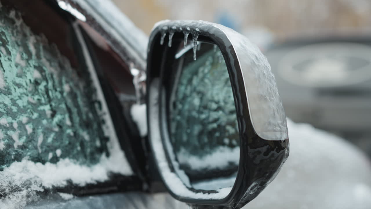 얼음으로 인 자동차 거울의 클로즈업, 자동차 창문의 반사와 함께, 겨울에 다른 눈으로 은 차의 흐릿한 배경을 보여줍니다.