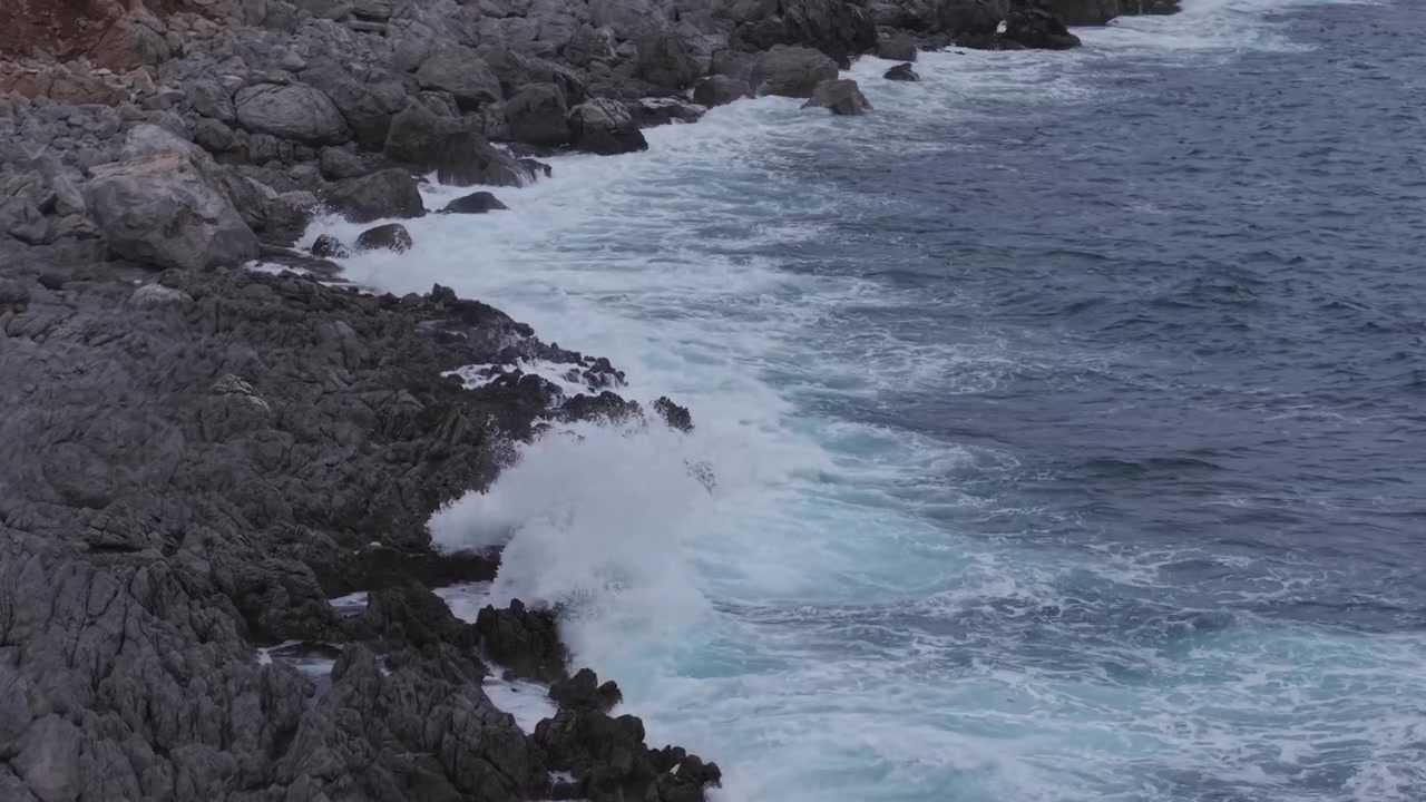 Forti onde oceaniche si infrangono su una costa rocciosa e frastagliata, creando schiuma bianca del mare e un movimento drammatico lungo la costa.
