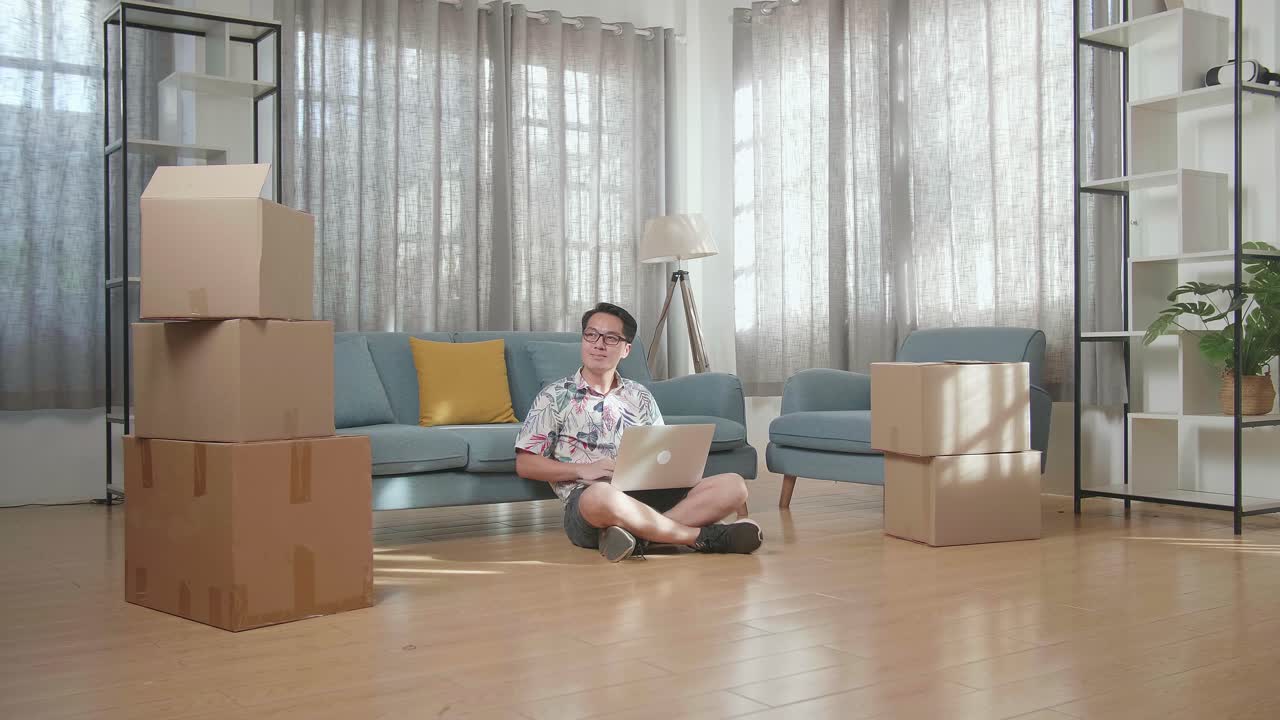 바닥에 앉아 새 집으로 이사한 후 노트북에 있는 사진과 집을 비교하는 카드보드 상자를 가진 아시아인