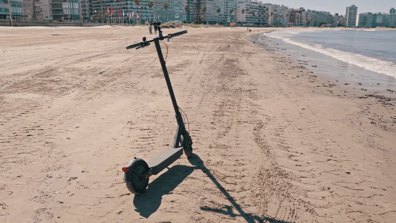 An electric scooter stands on a sandy beach with city buildings and the ocean in the background, perfect for travel and urban mobility concepts
