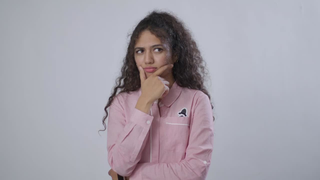 A South Asian woman with curly hair in a pink shirt stands with a thoughtful look, hand resting on chin, pondering