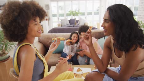 Happy-diverse-female-friends-sitting-on-sofa-in-living-room,-eating-pizza