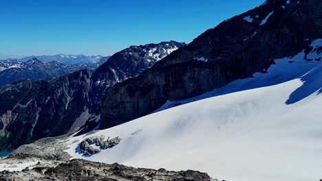 Summit-View-of-Joffre-Lakes-Provincial-Park-below-Climber-POV---Pan-Reveal-of-Turquoise-Water-from-Alpine-Snowy-Ridge-on-a-Sunny-Blue-Sky-Day