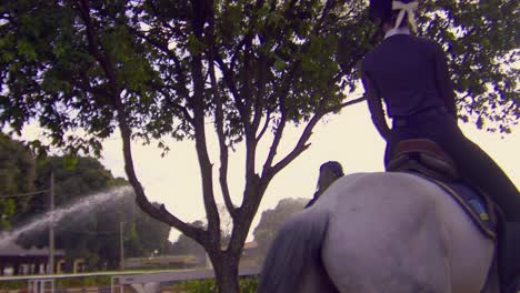 Cinematic-shot-of-a-beautiful-woman-riding-a-white-horse-while-wearing-a-black-and-white-outfit-on-a-beautiful-sunny-day-with-sprinklers-in-the-background