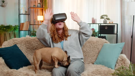 young woman using vr headset on home sofa with excited movement as pug dog watches with funny face