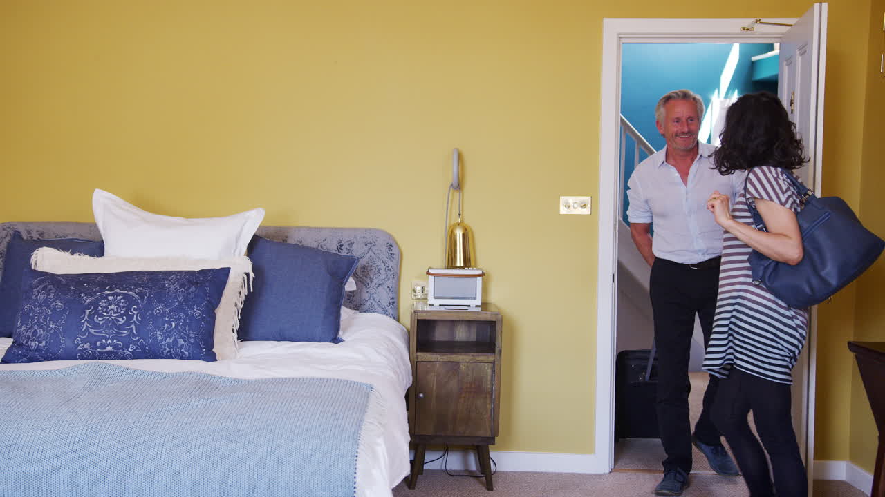 중년 부부가 도착하고 호텔 방을 감상하고, 포옹하고, 앞쪽을 바라보며, 방 안에서 볼 수 있습니다.