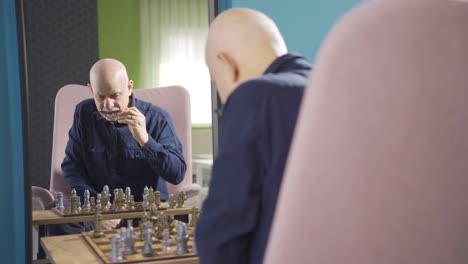 Focused-old-man-playing-chess-alone-at-home-and-looking-at-himself-in-mirror.