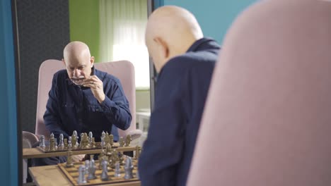 focused old man playing chess alone at home and looking at himself in mirror.