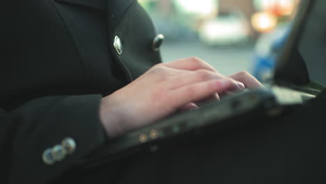 close up de un individuo escribiendo en una computadora portátil al aire libre mientras está vestido con un traje negro con botones plateados, el fondo borroso presenta coches estacionados y vegetación
