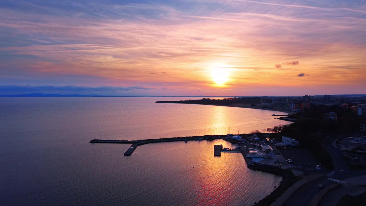 Kustlinje i skymningen, Bulgarien. Rosa och orange himmel som speglar sig i havet med vågbrytare, marina och avlägsen stadssiluett.