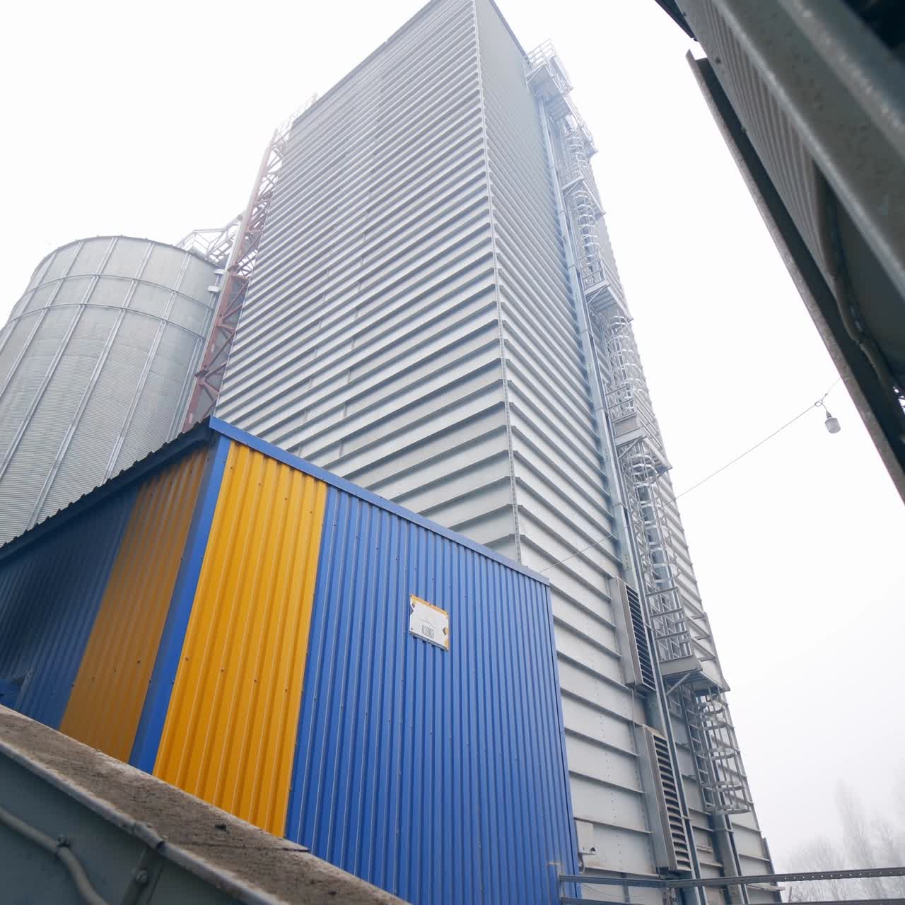 Huge silo tank with the metal ladder and a little balcony. A turning view from the grain elevator tank to other premises and cisterns in the agricultural plant