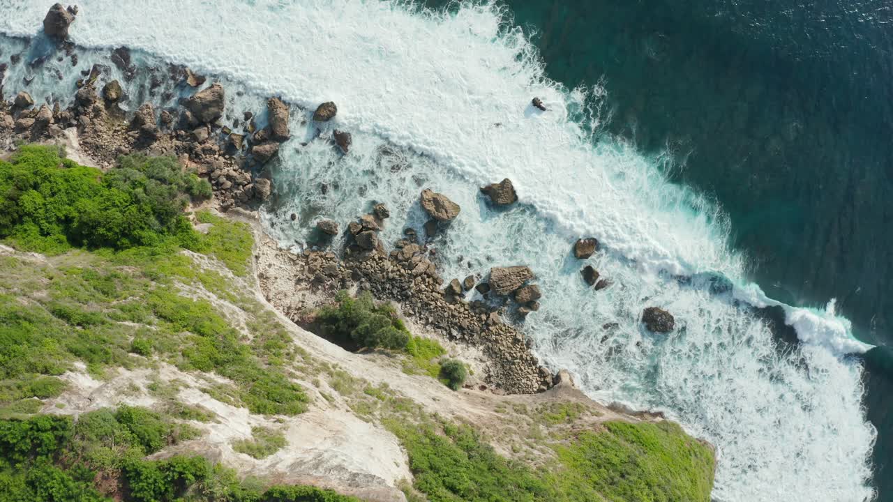 Aerial top down view of a rocky coastline with white foamy waves crashing into the shore