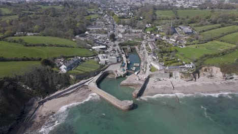 Aerial pan of stone-built harbour village where curved sea wall, docked boats and narrow lanes embrace a calm tidal inlet, with rolling green Cornish hills framing the coastline