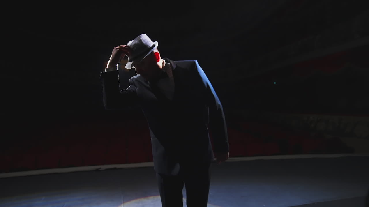 Male actor performing in the theater. Funny man putting on white hat and began dancing on stage in dark light. View from the stage.