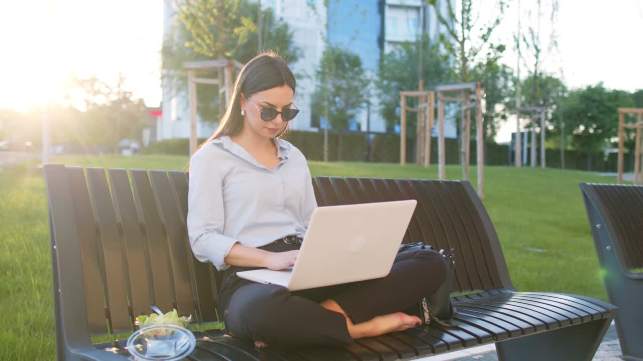 Business woman is sitting on bench outdoors and using lap top for working out office and eating salad