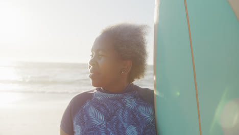 una mujer afroamericana mayor feliz sosteniendo una tabla de surf en la playa, en cámara lenta