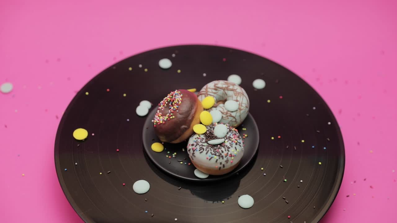 Two blue chocolate American donuts and one chocolate  donut roll together in a circle on a black and pink surface