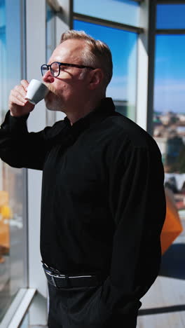 adult man stands at the window holding a cup in one hand, the other hand in the pocket. businessman drinking coffee in the office during a break from work. vertical video.