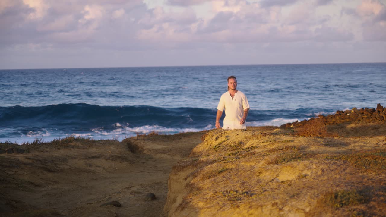 A man in white clothing stands still on a rugged coastal cliff, gazing thoughtfully at the vast blue ocean horizon under clear morning sunlight