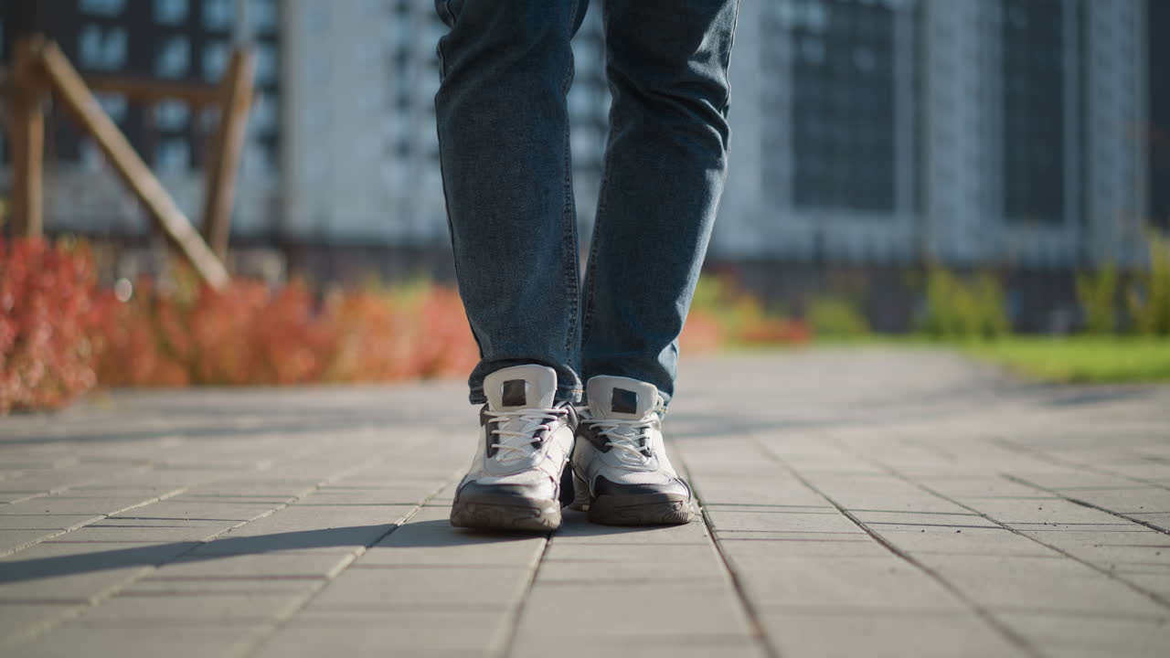Low-angle view of man in jeans and white canvas shoes walking slowly on interlocked pavement path with long shadow cast on ground during sunny day with blurred urban background