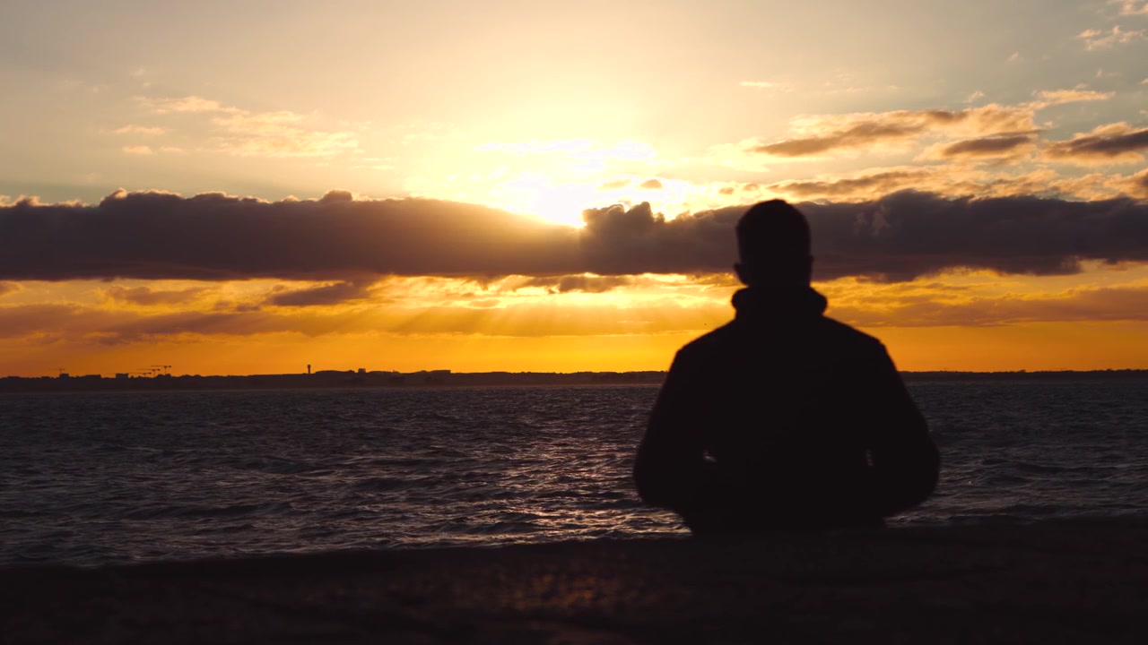 Silhouette di una persona che osserva un tramonto dorato su un mare agitato mentre le nuvole si muovono nel cielo serale