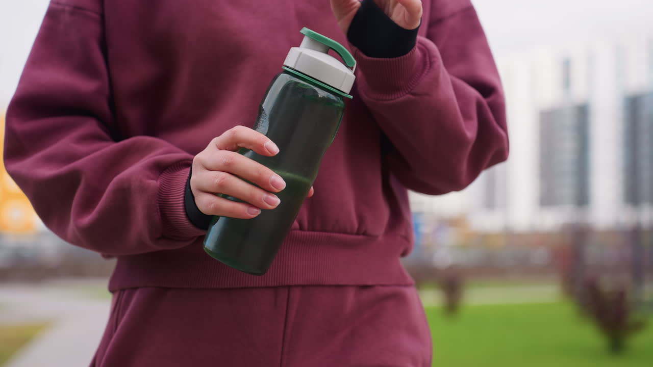 Lower body view of gym enthusiast in maroon hoodie grasping blue water bottle while walking along urban pavement with blurred building background, spring foliage, and distant city skyline