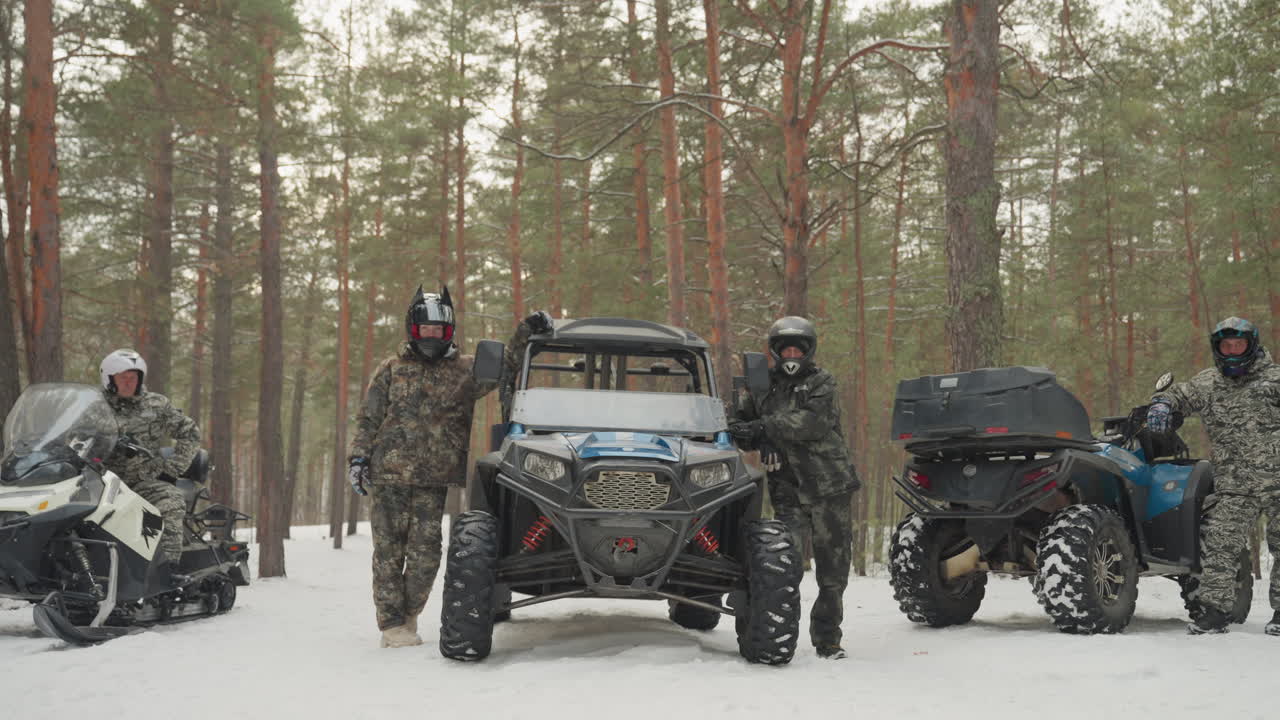 Tre piloti mimetici con quad e motoslitta in una pineta. Guide fuoristrada che preparano il percorso e controllano l'attrezzatura mentre sono in piedi accanto ai loro veicoli. Neve fresca sotto spessi tronchi, caschi e occhiali protettivi.