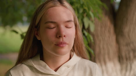 close-up of young lady closing and opening her eyes, basking in gentle sunlight with serene expression, enjoying soft breeze in natural setting, blurred green background with swaying leaves