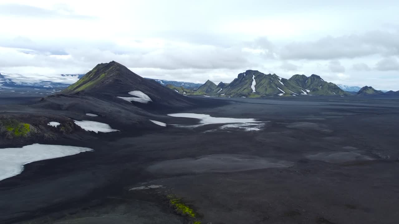 Aerial drone footage of dark black and brown green moss and white snow covered Iceland rocky volcanic terrain during a cloudy day with tall and big sharp edged mountains visible in the background.