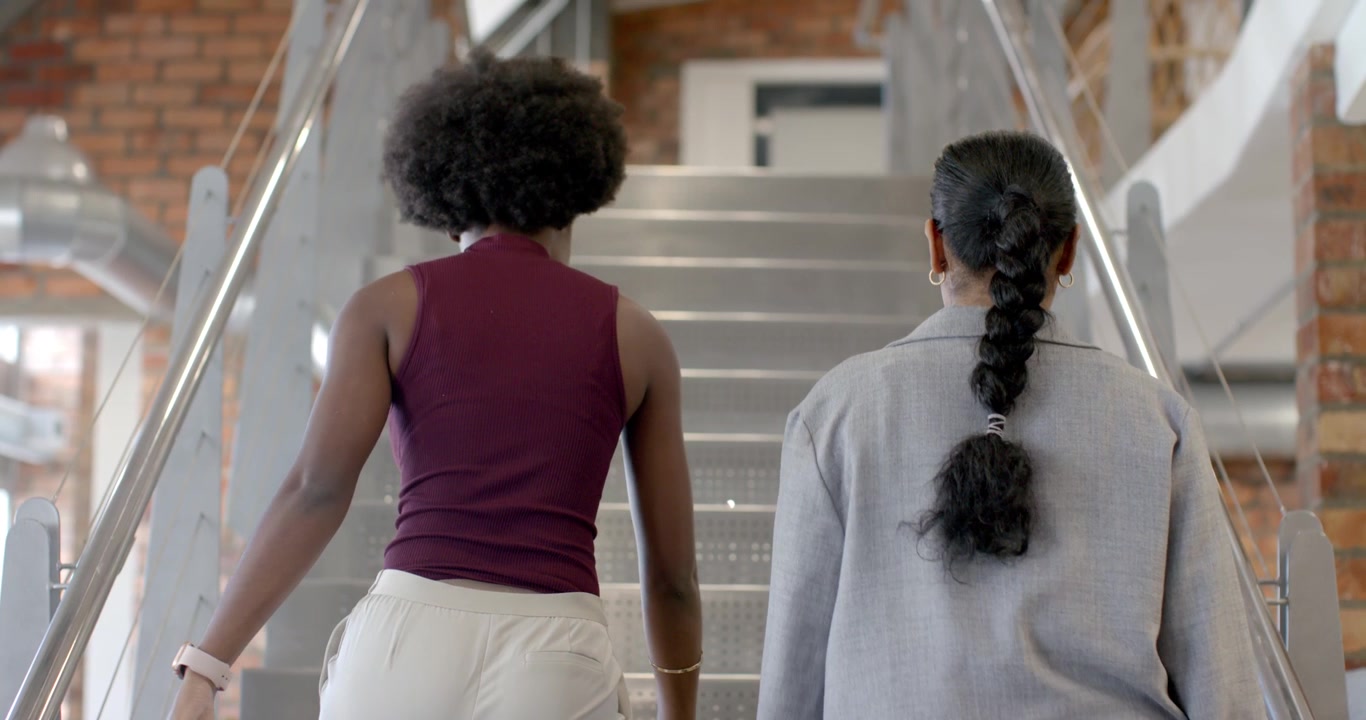 African American and Indian women climbing metal stairs carrying phone, joining on right heading up
