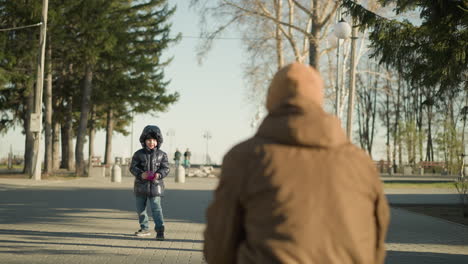 a close-up of a father and son playing outdoors, as the child releases a purple flying toy in the air the father rushes to catch it , with people walking in the background