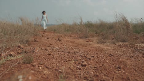 Low-angle-slow-motion-shot-of-a-dusty-and-dried-up-field-with-dry-plants-and-a-young-indian-woman-in-a-turquoise-dress-and-a-wood-basket-walking-to-the-camera-on-a-windy-day