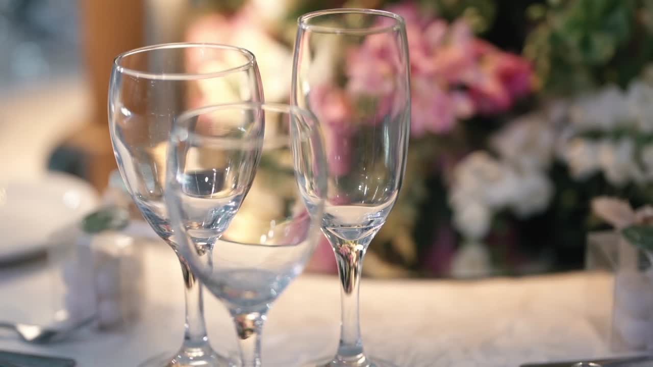 Dolly-in al rallentatore verso bicchieri vuoti su un tavolo elegantemente apparecchiato per un matrimonio, con fiori rosa tenue e bianchi sfocati sullo sfondo per creare un'atmosfera romantica e di lusso