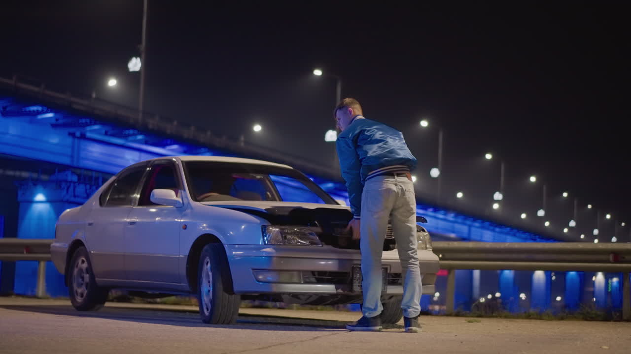 다리 아래에서 엔진을 점검하는 백인 운전자, 네온 블루 조명이 켜진 고가차도가 있는 야간 도시 풍경, 후드가 올라간 흰색 세단, 파란색 재킷과 청바지를 입고 엔진룸으로 몸을 기울인 남성, 가로등