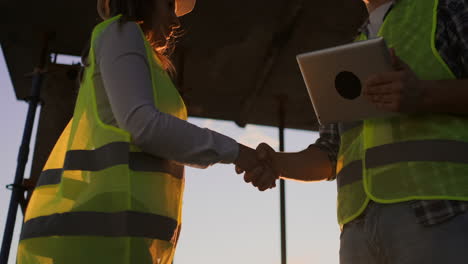 Ein-Bauarbeiter-Mit-Einem-Tablet-Und-Eine-Inspektorin-In-Weißen-Helmen-Geben-Sich-Bei-Sonnenuntergang-Die-Hand,-Stehen-Auf-Dem-Dach-Des-Gebäudes.-Symbol-Der-Zustimmung-Zu-Erfolgreicher-Arbeit