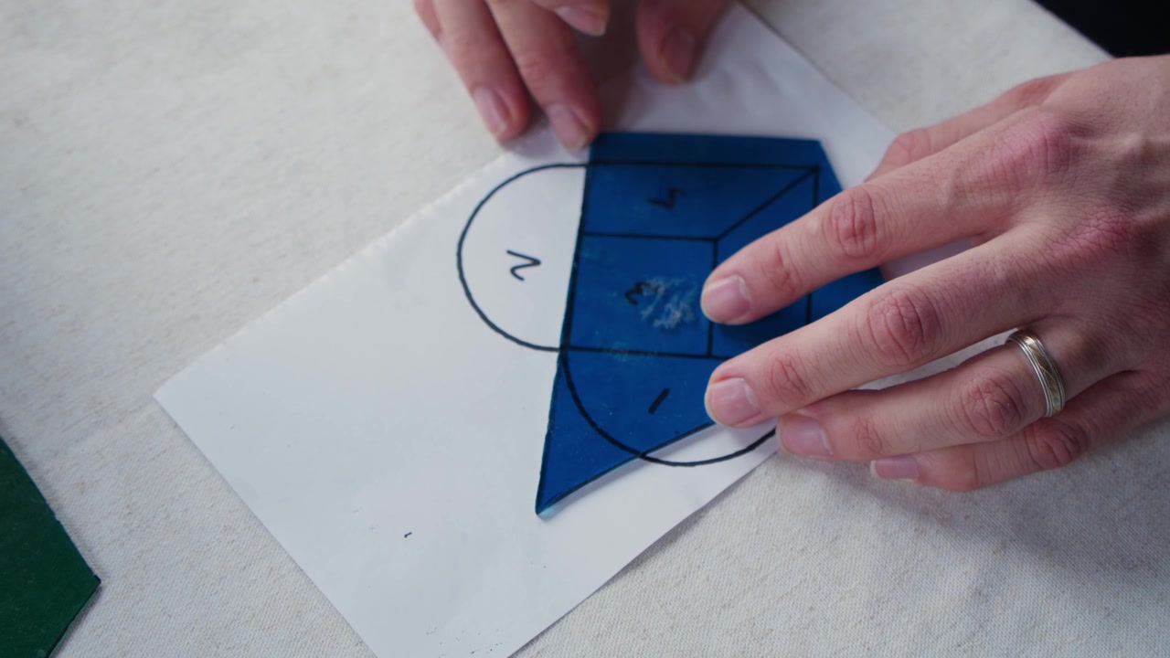 A man holds a blue piece of stained glass over a heart shaped template during an art class. The camera booms up to reveal more of the table.