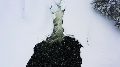 flying down towards dark pool at the bottom of a small waterfall as snowflakes fly upwards slow motion aerial