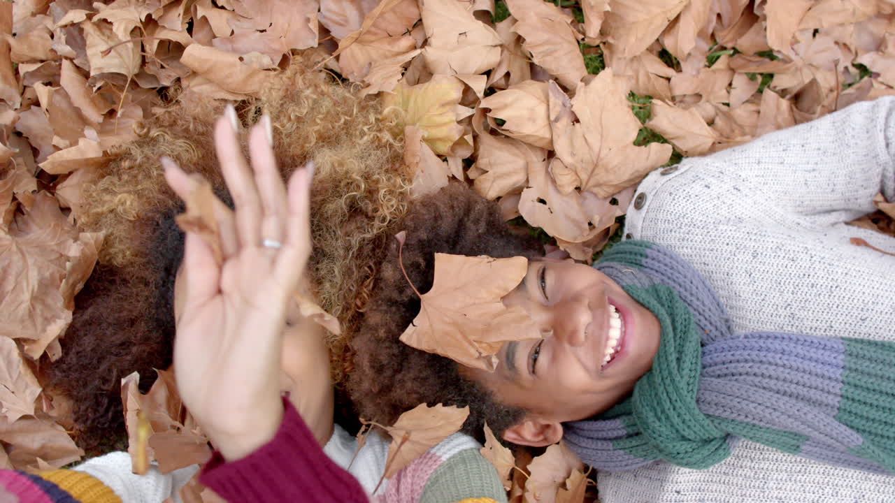 Happy african american mother and son in warm clothes lying on fallen leaves in garden, slow motion