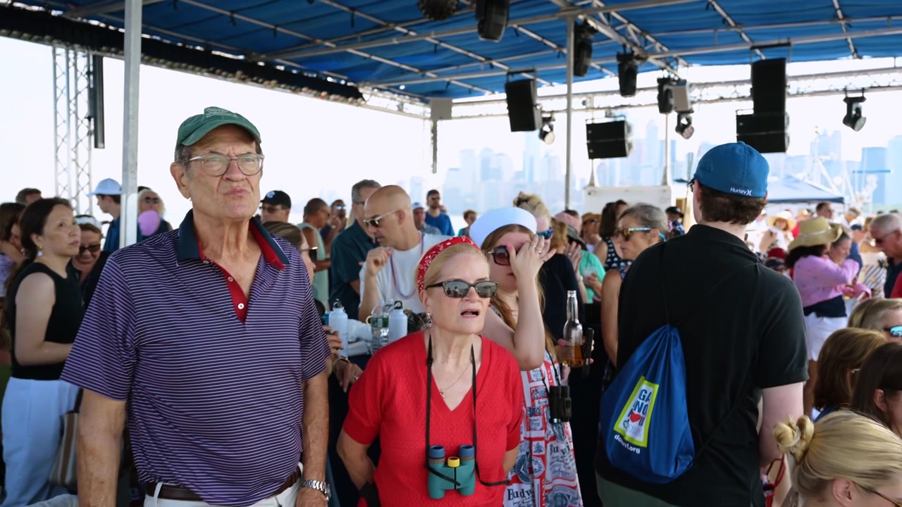 New York, USA, 4. juli 2026: Folk fejrer på et krydstogtskibs dæk. En menneskemængde af passagerer iført patriotisk tøj nyder en solrig bådtur med udsigt til en fjern bysilhuet