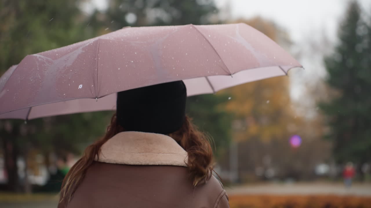 Thoughtful girl holding umbrella over head with light snowfall running outdoors wearing black knit cap and brown shearling jacket strands of hair blowing in breeze surrounded by autumn trees