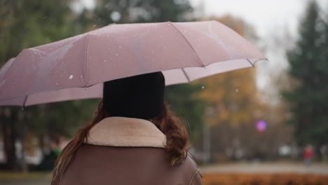 thoughtful girl holding umbrella over head with light snowfall running outdoors wearing black knit cap and brown shearling jacket strands of hair blowing in breeze surrounded by autumn trees
