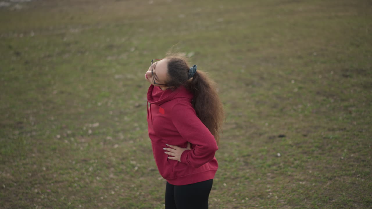 White Woman Performing Neck Mobility Stretches On Grass, Hands On Hips, Steady Breathing And Upward Rotations, Red Hoodie And Glasses, Focused Posture Work And Cool Open Field Setting