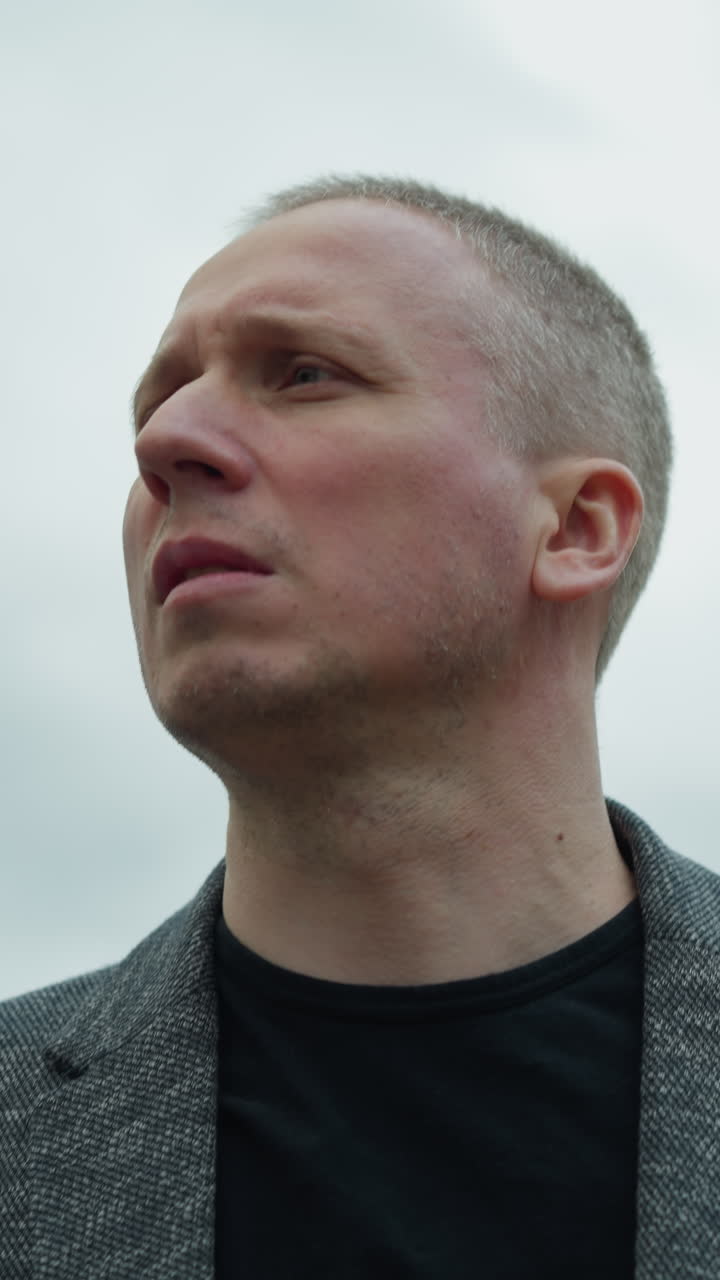 una vista de cerca de un hombre blanco envejecido con un traje gris y una camiseta polo negra, centrando su atención en el lado derecho, con un cielo nublado a la vista