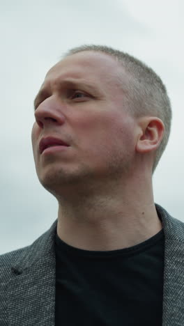 a close-up view of an aged white man wearing a grey suit and a black polo shirt, focusing his attention on the right hand side, with an overcast sky in view