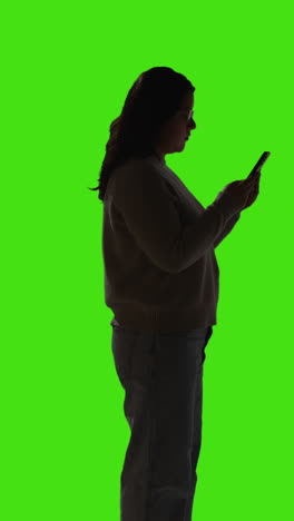 Vertical-Video-Silhouette-Shot-Of-Young-Woman-Using-Mobile-Phone-To-Text-Or-Check-Online-Standing-Against-Green-Screen