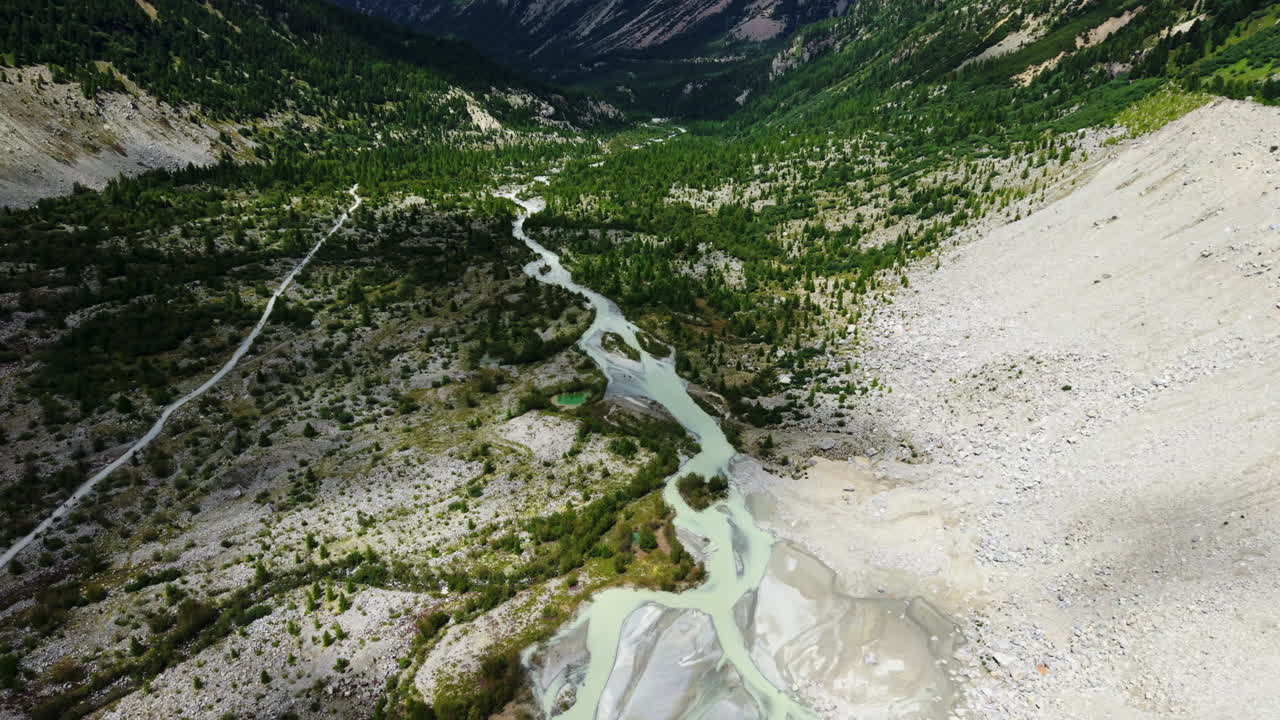 High drone shot showing the glacial river flowing through Morteratsch Valley in Switzerland, surrounded by rocky slopes and green forest