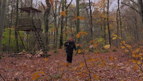 tiro largo de un cazador caminando lejos de una posición alta en un traje de invierno grueso, llevando un rifle de caza con un silenciador sobre su hombro